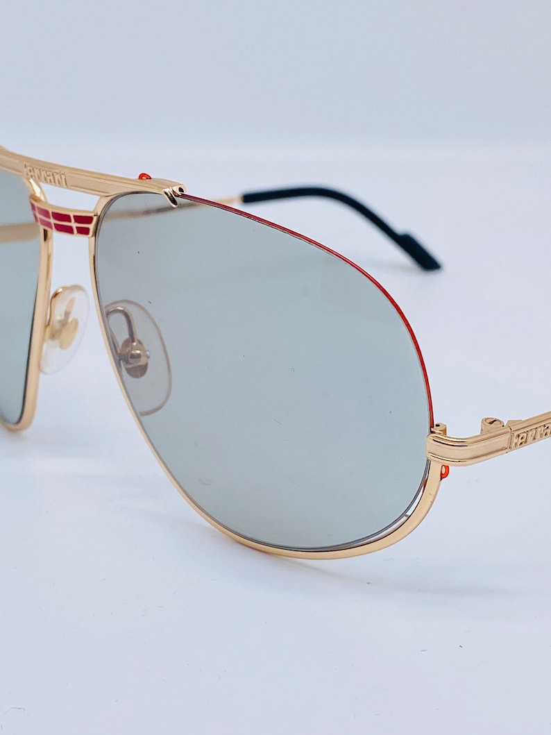 May include: Gold aviator sunglasses with a red stripe on the frame and gray lenses. The sunglasses have the word "Ferrari" on the side.