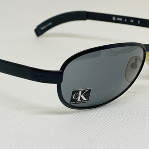 May include: Black oval-shaped sunglasses with gray lenses. The sunglasses have a thin black frame and arms. The Calvin Klein logo is visible on the lens and arm. The lenses provide 100% UV protection.
