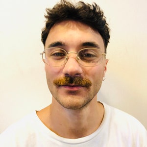 Może przedstawiać: Osoba nosząca okulary w złotej oprawie z widoczną na soczewce marką "GIORGIO ARMANI". Osoba ma na sobie biały t-shirt, wąsy i kr&oacute;tkie, ciemne włosy. Widoczny jest mały srebrny kolczyk w kształcie koła.