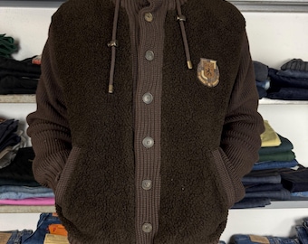 May include: A brown button-up jacket with a high collar and a textured, fuzzy body. The jacket features a knitted collar, sleeves, and trim, with a small emblem on the chest. The jacket has two pockets and is buttoned up.