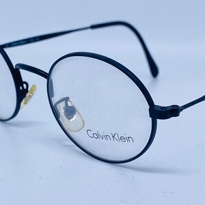 May include: Black round-framed eyeglasses with the brand name "Calvin Klein" printed on the right lens.