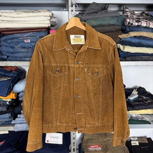 Pode incluir: Casaco de veludo cotelê castanho com fecho de botões, dois bolsos no peito e gola. O casaco está pendurado num cabide de madeira. A etiqueta diz "Slim Fit".