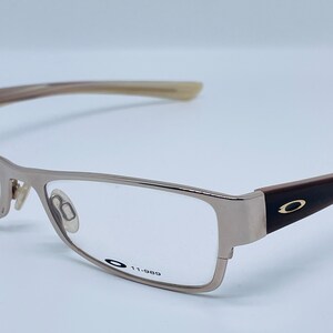 Może przedstawiać: Okulary prostokątne w kolorze srebrnym z brązowymi zausznikami i czarnym logo Oakley z boku. Okulary mają przezroczyste szkła, a na oprawce nadrukowany jest tekst "11-989".