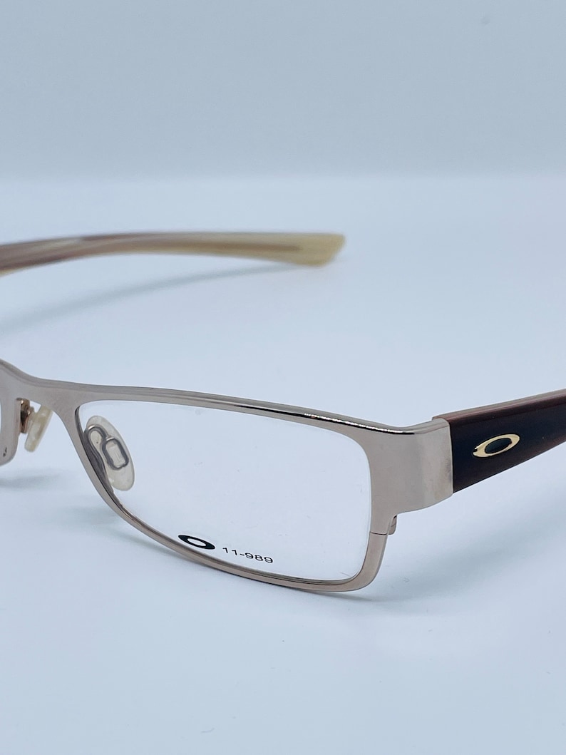 Może przedstawiać: Okulary prostokątne w kolorze srebrnym z brązowymi zausznikami i czarnym logo Oakley z boku. Okulary mają przezroczyste szkła, a na oprawce nadrukowany jest tekst "11-989".