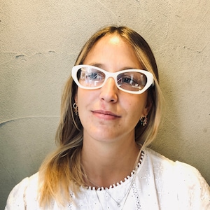 May include: A woman with blonde hair wearing white cat-eye glasses and a white lace top with a silver necklace and gold earrings.