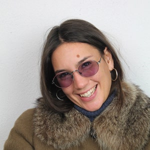Puede incluir: Una mujer con un abrigo marrón con cuello de piel y gafas redondas de color oscuro. Está sonriendo y mirando a la cámara.