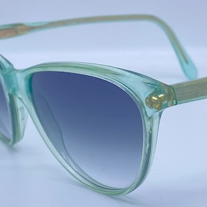 Puede incluir: Un par de gafas de sol de color verde claro con lentes azules degradados. Las gafas tienen un marco delgado y delicado.