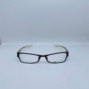 May include: Rectangular eyeglasses with a dark brown frame and clear lenses. The temples are a translucent light color. The glasses have a small logo on the lower right lens.
