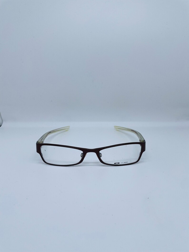 May include: Rectangular eyeglasses with a dark brown frame and clear lenses. The temples are a translucent light color. The glasses have a small logo on the lower right lens.