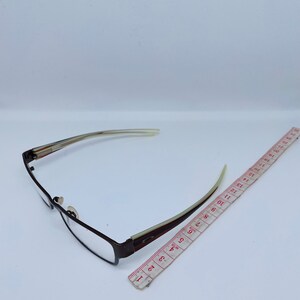 May include: Rectangular eyeglasses with a brown metal frame and clear lenses. The temples are a light beige color with brown and white stripes. A red and white measuring tape is next to the glasses.