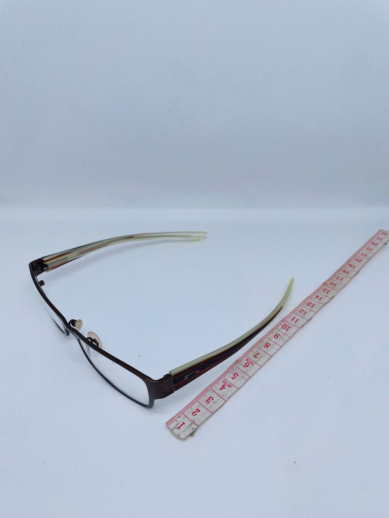 May include: Rectangular eyeglasses with a brown metal frame and clear lenses. The temples are a light beige color with brown and white stripes. A red and white measuring tape is next to the glasses.