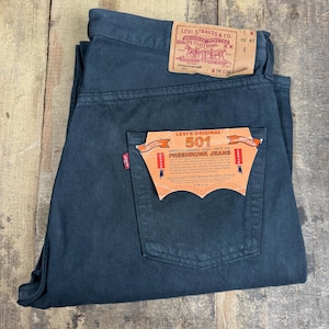 Può includere: Jeans Levi's 501 grigio antracite scuro con una toppa in similpelle beige in vita che riporta W38 L36. Una grande etichetta di carta arancione è attaccata alla tasca posteriore con il testo Levi's Original 501 Preshrunk Jeans e una piccola etichetta rossa.
