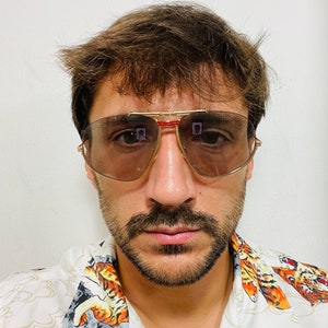 May include: A man with brown hair and a beard is wearing a white shirt with a tiger print and gold-rimmed sunglasses with brown lenses.