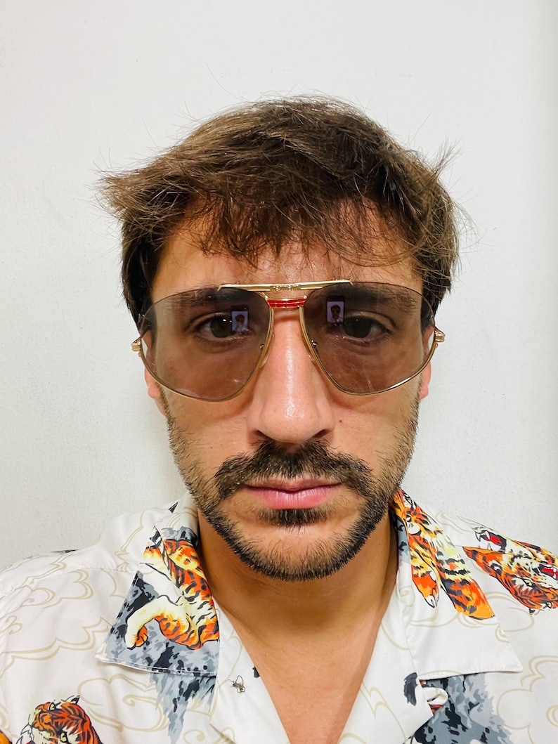 May include: A man with brown hair and a beard is wearing a white shirt with a tiger print and gold-rimmed sunglasses with brown lenses.