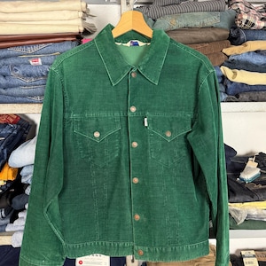 Pode incluir: Casaco de veludo cotelê verde com fecho de botões, dois bolsos no peito e gola. O casaco está pendurado num cabide de madeira. O casaco tem uma pequena etiqueta branca no bolso do peito. O casaco é exibido em frente a uma prateleira de jeans dobrados.