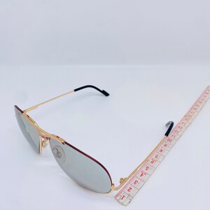 May include: Gold-tone aviator sunglasses with gray lenses and a red accent on the frame. The sunglasses are measured against a ruler.