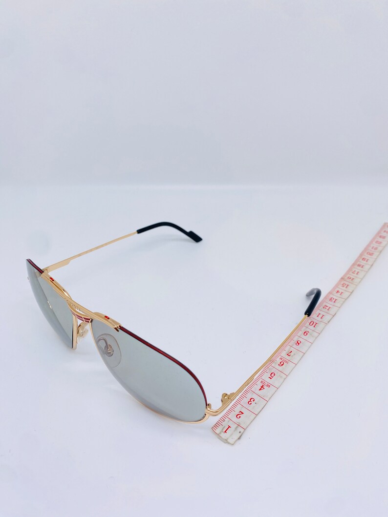 May include: Gold-tone aviator sunglasses with gray lenses and a red accent on the frame. The sunglasses are measured against a ruler.