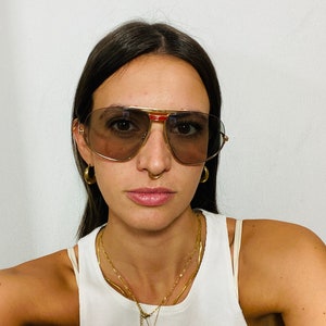 May include: A person wearing gold-rimmed aviator sunglasses with brown lenses and a red accent on the frame. They are wearing a white tank top and gold necklaces with a small octopus pendant.