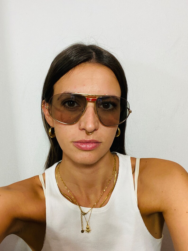 May include: A person wearing gold-rimmed aviator sunglasses with brown lenses and a red accent on the frame. They are wearing a white tank top and gold necklaces with a small octopus pendant.
