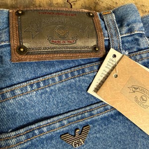 Puede incluir: Primer plano de unos vaqueros de mezclilla azules con un parche de cuero marrón y un emblema metálico. El parche dice "ARMANI JEANS" y "MADE IN ITALY". Una etiqueta de papel con el logo de Armani está adjunta.