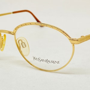 May include: Gold-tone eyeglasses with oval lenses. The frames feature a delicate design along the top rim and temples. The brand name "Yves Saint Laurent" is printed on the lens. The glasses have a classic, elegant style.