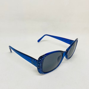 Puede incluir: Gafas de sol de pl&aacute;stico azul con pedrer&iacute;a alrededor de las monturas. Las lentes son de color gris oscuro.