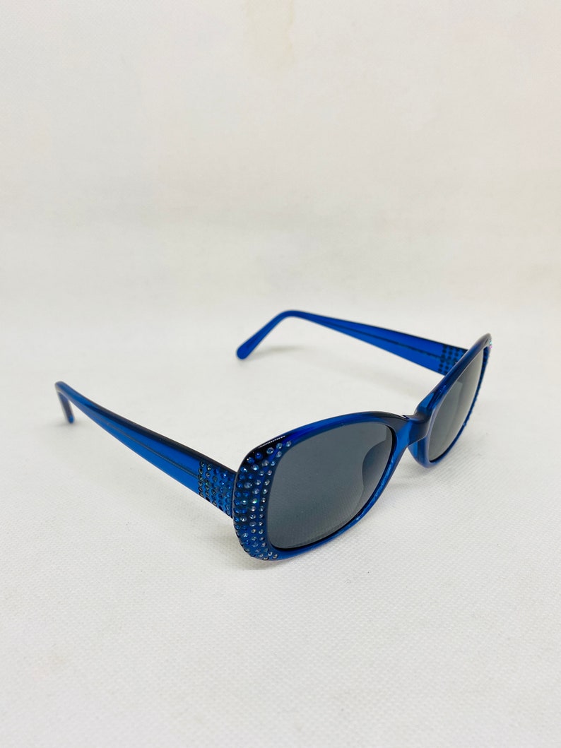 Puede incluir: Gafas de sol de pl&aacute;stico azul con pedrer&iacute;a alrededor de las monturas. Las lentes son de color gris oscuro.