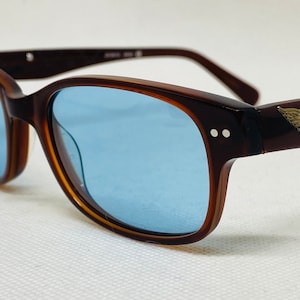 Può includere: Occhiali da sole con montatura marrone tartaruga e lenti blu. Gli occhiali hanno un piccolo logo dorato sulla stanghetta destra.
