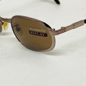 Op de afbeelding: Bruine metalen zonnebril met bruine glazen. De zonnebril heeft een label met de tekst "100% UV Protection Replay".