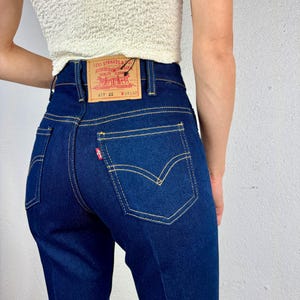 Może przedstawiać: Dżinsy z ciemnego denimu o wysokim stanie i prostym kroju nogawek. Na tylnej kieszeni znajduje się metka Levi's z napisem "Levi Strauss & Co. Quality Clothing" i rozmiarem "W 28 L 32".