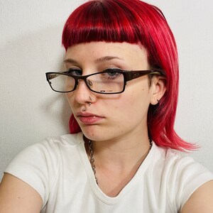May include: A person with bright red hair and glasses. They are wearing a white t-shirt with the text "Radio Silence" in a black box. A silver chain necklace is visible.
