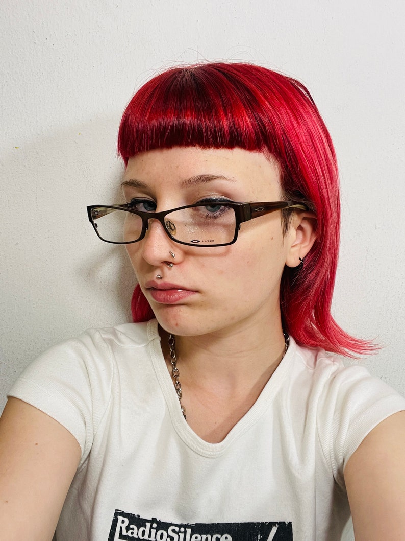 May include: A person with bright red hair and glasses. They are wearing a white t-shirt with the text "Radio Silence" in a black box. A silver chain necklace is visible.