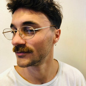 Może przedstawiać: Okrągłe okulary w złotej oprawie z nazwą marki "GIORGIO ARMANI" wydrukowaną na soczewce. Okulary nosi osoba z wąsami i białą koszulką.