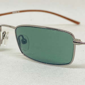 May include: Silver framed sunglasses with green lenses. The sunglasses have a thin, brown metal frame.