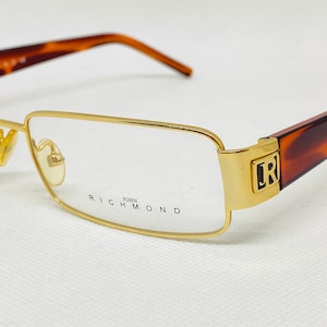 Puede incluir: Gafas de montura dorada con patillas de carey marrón. Las gafas tienen el logotipo de John Richmond en la patilla derecha y el nombre de la marca en la patilla izquierda.