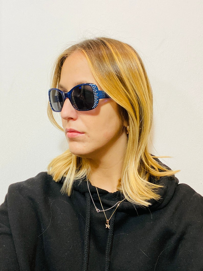Puede incluir: Una mujer con una sudadera con capucha negra, un collar de estrella dorada y gafas de sol azules con pedrer&iacute;a.