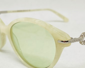 Puede incluir: Gafas de sol redondas blancas con lentes verdes y montura de metal plateado. La montura tiene un adorno de cristal en el lateral con la palabra "SWAROVSKI" impresa.