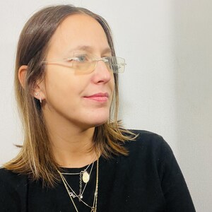 May include: A woman with brown hair wearing clear framed glasses and a black shirt with multiple gold necklaces.