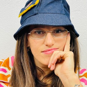 May include: A woman wearing a blue denim bucket hat with a yellow patch, glasses, and a brown and pink patterned shirt. She is wearing a beaded bracelet with an anchor charm on her wrist.