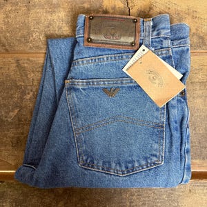 Puede incluir: Vaqueros de mezclilla azul claro con un parche de cuero marrón y una etiqueta. Los vaqueros presentan un diseño clásico de cinco bolsillos y el logo de Armani en el bolsillo trasero. Los vaqueros están doblados sobre una superficie de madera.