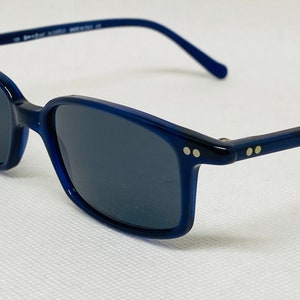 May include: Blue framed sunglasses with dark lenses. The frames have a subtle curve and two small gold dots on each side.