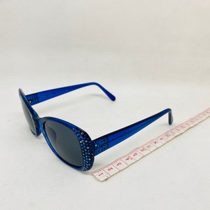 Puede incluir: Gafas de sol ovaladas azules con pedrer&iacute;a alrededor del marco. Las gafas de sol est&aacute;n sobre una superficie blanca con una cinta m&eacute;trica.
