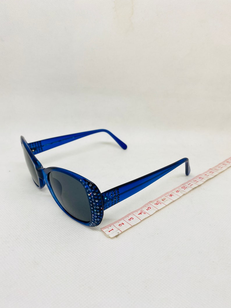 Puede incluir: Gafas de sol ovaladas azules con pedrer&iacute;a alrededor del marco. Las gafas de sol est&aacute;n sobre una superficie blanca con una cinta m&eacute;trica.