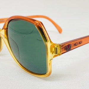 May include: Orange and brown plastic sunglasses with green lenses. The sunglasses have a geometric shape and a logo on the side.