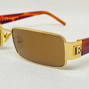 Puede incluir: Gafas de sol doradas con lentes marrones y patillas de carey. Las gafas tienen una montura rectangular y la patilla derecha tiene la letra "R" en oro.