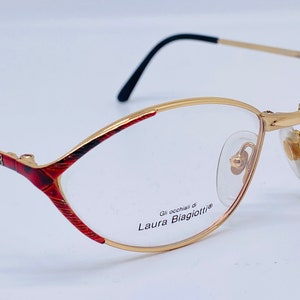 May include: Gold-rimmed eyeglasses with a red and black patterned frame. The glasses have the text "Gli occhiali di Laura Biagiotti®" printed on the frame.
