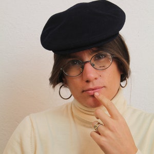Puede incluir: Una persona con una gorra de marinero azul marino, gafas redondas y un jersey de cuello alto crema. Lleva un anillo de oro en el dedo y tiene el dedo en la barbilla.