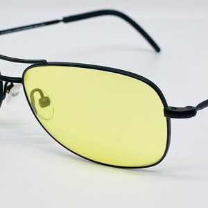 Puede incluir: Gafas de sol aviador negras con lentes amarillos. Las gafas tienen un fino marco de metal y están diseñadas para uso al aire libre.