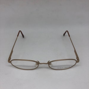 Może przedstawiać: Złote okulary Giorgio Armani z owalnymi soczewkami i smukłymi zausznikami. Oprawki mają minimalistyczny design z brązowymi końc&oacute;wkami zausznik&oacute;w. Nazwa marki jest wydrukowana na prawym dolnym szkle.