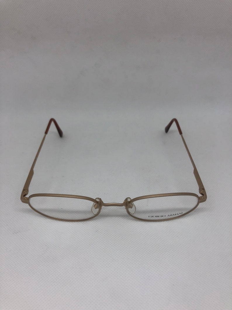 Może przedstawiać: Złote okulary Giorgio Armani z owalnymi soczewkami i smukłymi zausznikami. Oprawki mają minimalistyczny design z brązowymi końc&oacute;wkami zausznik&oacute;w. Nazwa marki jest wydrukowana na prawym dolnym szkle.